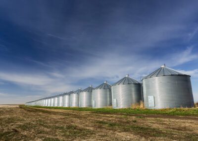A Long Row Of Shiny Metal Grain Bins Reflecting Sunlight With Blue Sky And Clouds